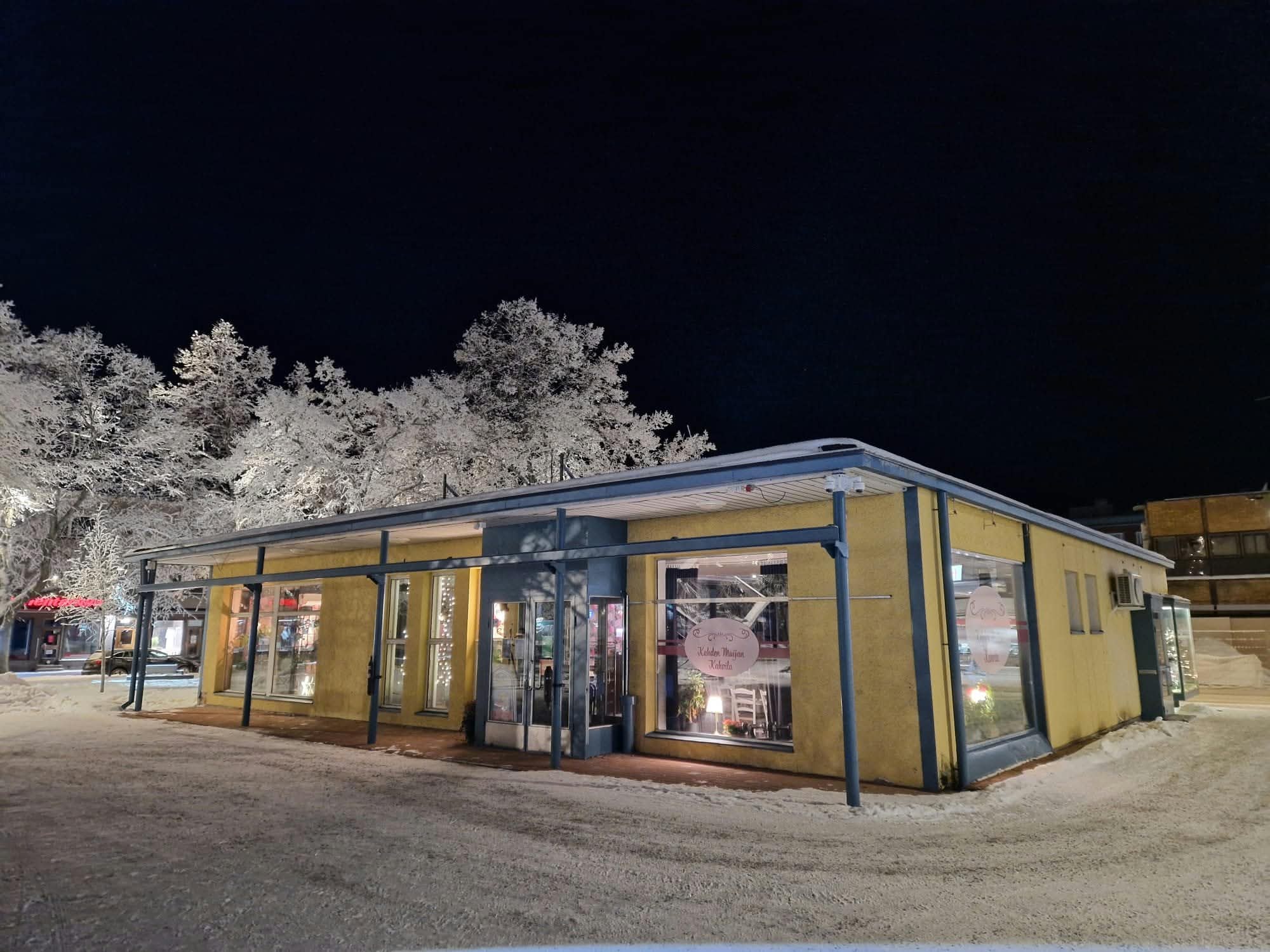 Kahvilan keltainen julkisivu sinisine pilareineen pimeänä talviyönä, ikkunoista välähtää lämmin valo ja maassa on lunta.